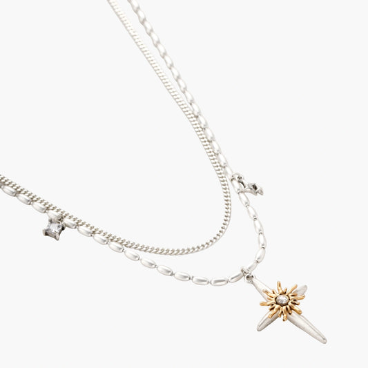 Layered silver-toned necklace set with cross pendant and gold sun charm detail, matching drop earrings on white background.